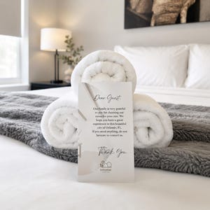 Puede incluir: Una escena de habitación de hotel con toallas blancas enrolladas y una tarjeta de bienvenida. La tarjeta dice "Estimado huésped" y expresa gratitud por elegir la habitación. La habitación tiene una manta gris y una lámpara.