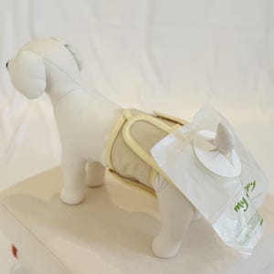 Può includere: Un pannolino beige per cani con rifiniture gialle è indossato da un cane di peluche. Un sacchetto di plastica trasparente con le parole "my puppy" in verde è attaccato al pannolino. Il cane è su una superficie bianca.