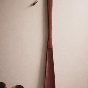 May include: A long, wooden shoe horn with a dark reddish-brown finish. It has a slender handle and a curved end for easy shoe wear. A leather loop is attached for hanging. The shoe horn is approximately 20 inches long.