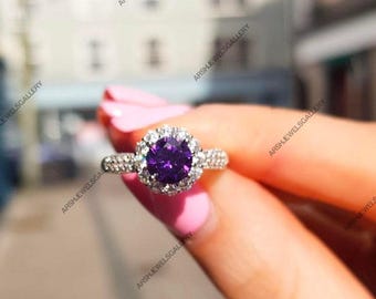 May include: A silver ring with a large, round, deep purple gemstone at the center, surrounded by a halo of smaller, clear stones. The ring's band is also adorned with small, clear stones, creating a sparkling effect. The ring is held up by a person.