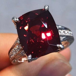 Puede incluir: Un anillo plateado con una gran gema roja oscura tallada en cojín. La gema facetada captura la luz y refleja un tono rojo brillante. La banda está adornada con gemas más pequeñas y claras.