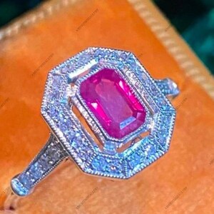 Puede incluir: Primer plano de un anillo plateado con una gema rosa rectangular y vibrante en el centro. La gema está rodeada por un halo de piedras más pequeñas y transparentes. El anillo tiene un diseño intrincado y detallado, de estilo vintage.