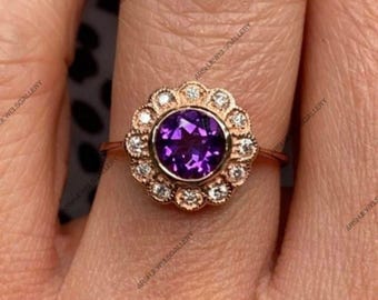 May include: Rose gold ring featuring a large, round, deep purple gemstone at the center, surrounded by a halo of smaller, clear gemstones. The ring's setting has a floral design. The ring is on a finger.