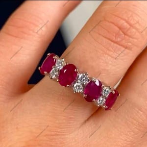 Puede incluir: Un anillo de plata con gemas de rubí de corte ovalado que se alternan con gemas claras redondas más pequeñas. Los rubíes son de color rojo intenso y el anillo está en un dedo.