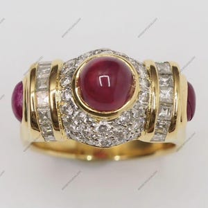 Può includere: Un anello d'oro con un grande rubino rotondo rosso scuro al centro, circondato da diamanti più piccoli. Due rubini aggiuntivi sono incastonati ai lati. L'anello presenta un design classico ed elegante.