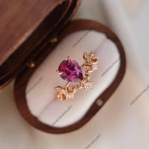 Puede incluir: Un anillo de oro rosa con una piedra preciosa rosa oscuro en forma de pera. El anillo tiene un diseño floral con pequeñas piedras de acento transparentes. El anillo se muestra dentro de un joyero de madera.
