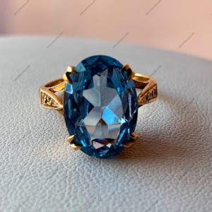 Könnte beinhalten: Ein goldener Ring mit einem großen, oval geschliffenen, blauen Edelstein. Der Edelstein ist der Mittelpunkt, gefasst in einem goldenen Band mit dekorativen Filigranarbeiten an den Seiten. Der Ring wird auf einer hellen Oberfläche präsentiert.