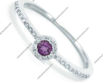 May include: A silver ring with a central, round, purple gemstone surrounded by a halo of smaller, clear stones. The band is also adorned with small, clear stones, creating a sparkling effect. The ring is set against a white background.