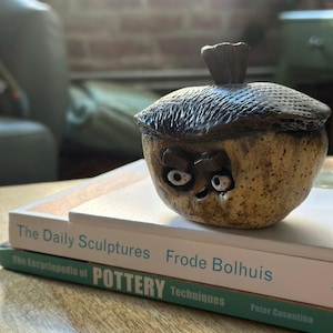 May include: A handcrafted ceramic acorn-shaped container with a lid. The container is tan with dark brown accents and has a whimsical face carved into the front. The lid has a textured surface and a small stem. The container is sitting on top of two books.