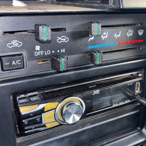 May include: Close-up of a car's dashboard featuring a JVC stereo and climate control panel. The panel has black knobs with green stripes, and the stereo has a silver knob and yellow buttons. The A/C button is visible.