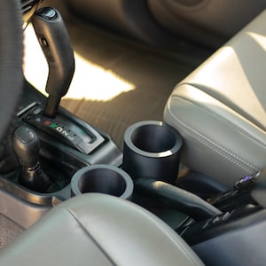 May include: A black cylindrical cup holder insert positioned in the center console of a vehicle, situated between the gear shift lever and the handbrake, featuring a smooth matte finish and a deep, hollow interior designed for holding beverage containers.