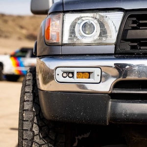 May include: A close-up of a grey truck's front, highlighting a headlight and bumper with a white light housing. The bumper has a chrome finish. The truck's tyre is visible on the left, and a vehicle with colourful stripes is in the background.