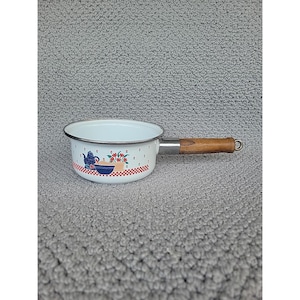 May include: White enamel saucepan with a wooden handle. The pan features a decorative pattern of a blue teapot, red flowers, and a blue bowl on a red and white checkered border. The rim is trimmed with a silver band.