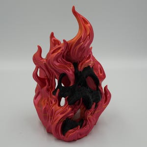 Könnte beinhalten: Eine 3D-gedruckte Skulptur einer stilisierten Flamme mit einem schädelartigen Gesicht. Die Flamme ist hauptsächlich rot mit orangefarbenen Akzenten, das Gesicht ist schwarz. Die Skulptur hat eine glatte, glänzende Oberfläche.