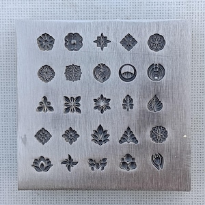 May include: A silver metal stamping block with various floral and leaf designs. The block features an array of detailed, engraved patterns, including flowers, leaves, and geometric shapes. The designs are arranged in neat rows and columns, showcasing a variety of botanical motifs.