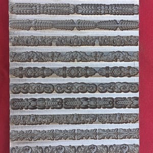 May include: A rectangular metal plate with multiple rows of decorative border designs. Each row features a different embossed pattern, including floral, scroll, and geometric motifs. The plate is set against a red background.