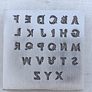 May include: A square metal plate with the alphabet engraved in a decorative font. The letters are arranged in rows and columns, with each letter having a unique, ornate design. The plate has a brushed metal finish.