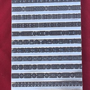 May include: A rectangular metal plate with multiple rows of decorative border designs. Each row features a different embossed pattern, including floral, geometric, and stylized motifs. The plate is silver-colored, and the designs are dark gray.