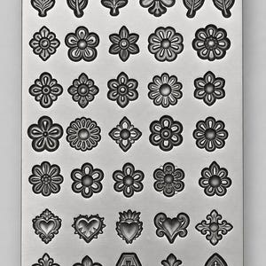 May include: A rectangular metal mold featuring a grid of thirty-five recessed cavities. The cavities are shaped as various floral patterns, leaves, hearts, fleur-de-lis symbols, a coffin, and a gothic arch window with a cross design.