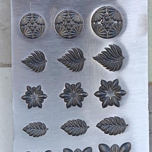 Puede incluir: Placa de metal con varios diseños florales y de hojas en relieve. Los diseños incluyen girasoles, hojas y formas de flores estilizadas. El metal tiene un acabado cepillado y los diseños son de un color más oscuro.