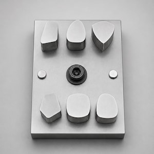 Pode incluir: Uma placa metálica retangular com seis formas geométricas elevadas de bordos lisos, dispostas em duas filas de três. Um parafuso circular preto com encaixe hexagonal está posicionado ao centro, ladeado por dois pequenos rebites metálicos planos.