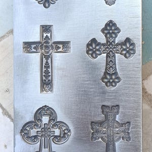 Könnte beinhalten: Silberne Metallplatte mit sechs geprägten Kreuzen in verschiedenen Designs. Jedes Kreuz weist ein einzigartiges, verziertes Muster mit floralen Elementen und filigranen Linien auf. Die Platte ist rechteckig.