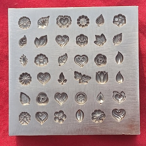 Puede incluir: Un bloque de estampado de metal para joyería con varios diseños florales y de hojas. Los diseños incluyen flores, corazones y hojas, todos en relieve. El bloque es rectangular y tiene un acabado de metal cepillado.