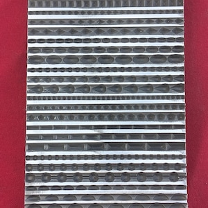 May include: A rectangular metal tool with various textured patterns. The patterns include horizontal lines, circles, and geometric shapes. The tool is silver and sits against a red background.