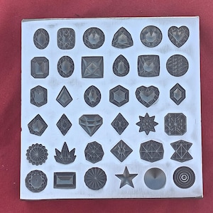 Puede incluir: Una placa de metal plateado con 36 formas de piedras preciosas grabadas, incluyendo cuadrados, círculos, corazones y estrellas. Cada forma presenta grabados detallados, dando una apariencia texturizada. La placa se encuentra sobre un fondo rojo.