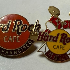 Könnte beinhalten: Zwei Hard Rock Cafe Pins. Ein Pin zeigt den Text "Hard Rock Cafe San Francisco" in goldfarbenen Buchstaben auf rotem Hintergrund. Der andere Pin hat ein Schlagzeug-Design mit dem Text "Hard Rock Cafe Los Angeles".