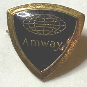 Könnte beinhalten: Eine Vintage-Amway-Anstecknadel mit goldfarbenem Rand und schwarzem Mittelteil. Die Anstecknadel zeigt ein goldfarbenes Weltkugel-Design und das Wort "Amway" in goldfarbenen Buchstaben. Die Anstecknadel ist schildförmig.