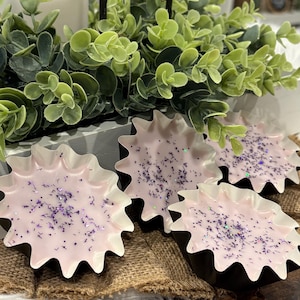 Op de afbeelding: Vier stervormige wax melts met een lichtroze kleur en paarse glitter. De melts liggen op een jute oppervlak. Een witte kaars en groen gebladerte zijn op de achtergrond.
