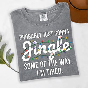A gray t-shirt with a humorous text printed on it, along with a brown hat and some greenery in the background.