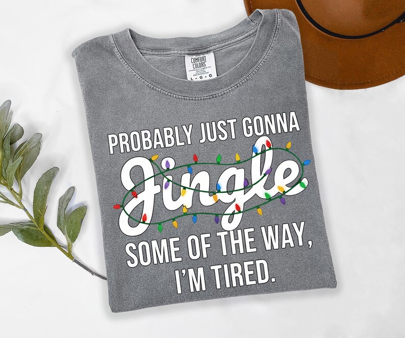 A gray t-shirt with a humorous text printed on it, along with a brown hat and some greenery in the background.