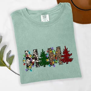 Może przedstawiać: Jasnozielona koszulka Comfort Colors z napisem "MAMA" w świątecznym wzorze. Litery zdobione są nadrukiem krowim, lamparcim, kratą i czerwonym wzorem w kratę bawolą, z lampkami choinkowymi.