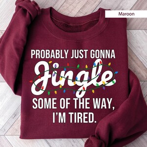 A maroon sweatshirt with a humorous text printed on it, which reads "Probably Just Gonna Jingle Some of the Way, I'm Tired."