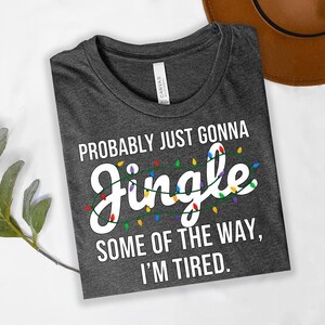 A gray t-shirt with a humorous text printed on it, lying on a white surface next to a brown hat.