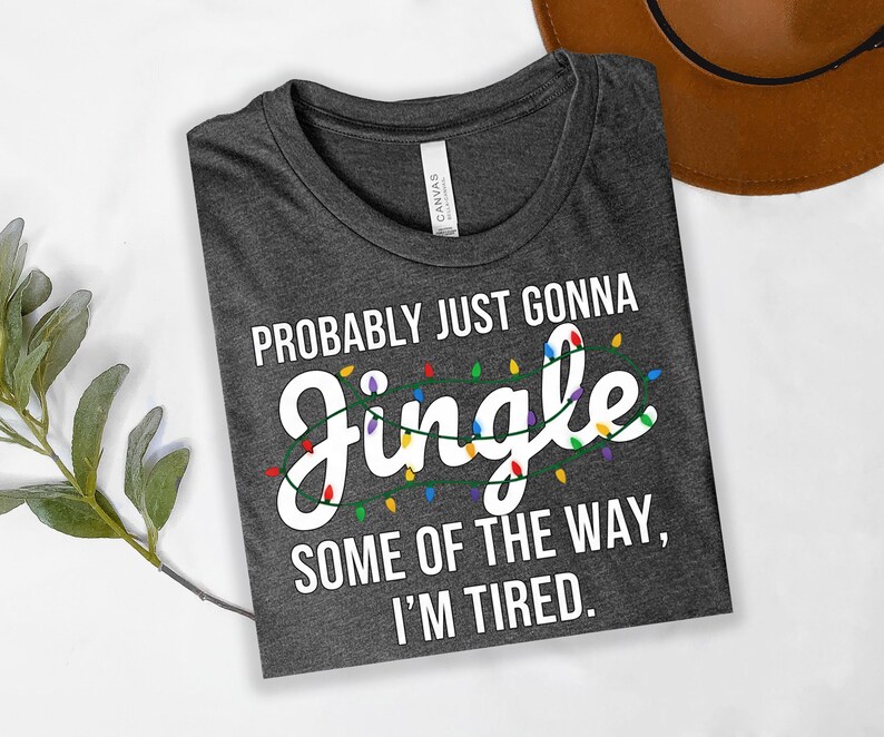 A gray t-shirt with a humorous text printed on it, lying on a white surface next to a brown hat.