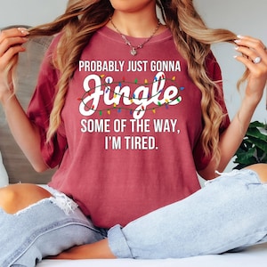 A woman wearing a red t-shirt with text and a necklace is sitting on a bed with her hair in a ponytail.