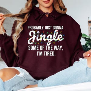 A woman wearing a burgundy hoodie with a humorous text printed on it, sitting on a bed with her legs crossed.