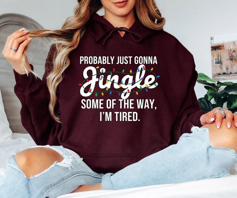 A woman wearing a burgundy hoodie with a humorous text printed on it, sitting on a bed with her legs crossed.