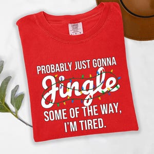 A red t-shirt with a humorous text printed on it, lying on a white surface next to a brown hat.