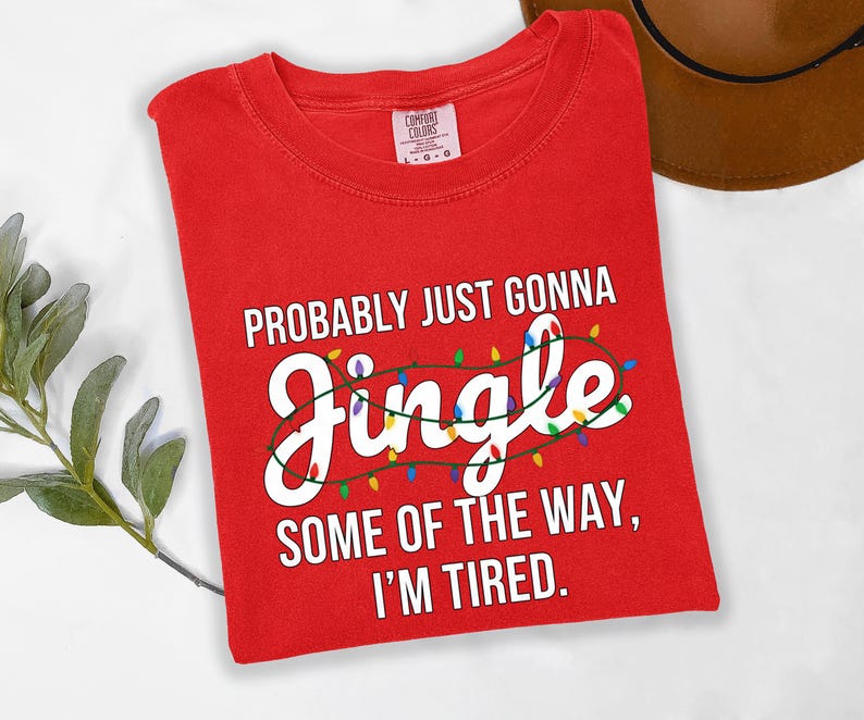 A red t-shirt with a humorous text printed on it, lying on a white surface next to a brown hat.