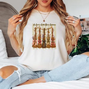 May include: A cream-colored t-shirt featuring a graphic print of five decorative broomsticks adorned with flowers, ribbons, and charms. Above the brooms, the text reads A Girl's Gotta Have Options. The design is set against a background of vertical stripes.