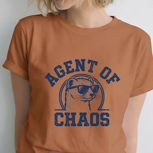 May include: A terracotta-coloured t-shirt with rolled sleeves. The shirt displays a navy blue graphic of a weasel wearing sunglasses, with the words "AGENT OF CHAOS" in a circle.