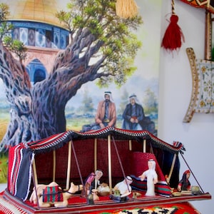 Puede incluir: Una escena en miniatura de una tienda de campaña de Oriente Medio con figuras y un camello, colocada sobre una alfombra roja estampada. La tienda tiene un techo a rayas rojas y negras. Una pintura de un edificio con una cúpula dorada y un gran árbol está en el fondo.