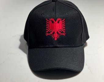 May include: Black baseball cap with a red embroidered double-headed eagle emblem. The cap has a curved brim and a button on top. The eagle design is centered on the front of the cap.