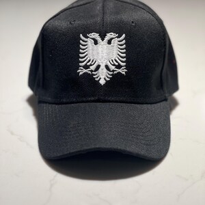 Może przedstawiać: Czarna czapka bejsbolówka z białym haftowanym emblematem dwugłowego orła. Czapka ma zakrzywiony daszek i guzik na górze. Kapelusz jest wykonany z teksturowanej tkaniny.