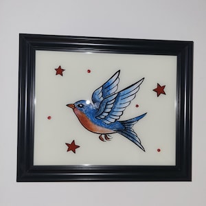 May include: Framed artwork featuring a bluebird with outstretched wings, accented with orange and black details. The bird is surrounded by red stars and dots on a cream-colored background, all encased in a black frame.