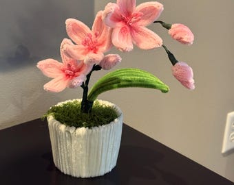 May include: A decorative artificial orchid plant in a white textured pot. The plant features pink flowers, green leaves, and a mossy base. The flowers have a detailed, textured appearance, and the pot has a ribbed design.